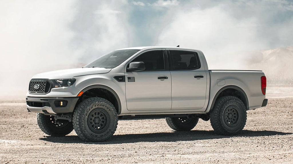 The silver Ford Ranger truck is driving on a dirt road under a cloudy sky.