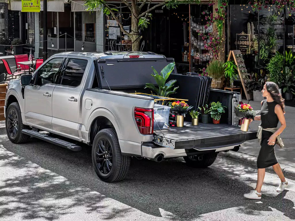 A silver pickup truck with an open tailgate loaded with plants on a city street 