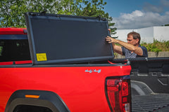 Man adjusting a black tonneau cover on a red truck bed with a white building and trees in the background.